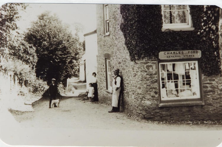 Charles Ford leaning against the wall of his shop in Kellaton. The little girl with bare feet is Ceda Ford, the woman with her is probably Florrie Harradon, daughter of the local blacksmith Charlie Harradon. The boy is Bill Wright child a local boy who was apprenticed to and lived with the Harradons. Date is probably 1911.